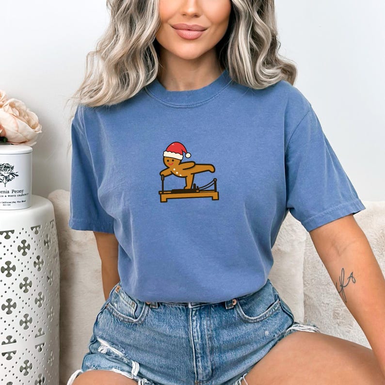 May include: A blue t-shirt featuring a gingerbread man wearing a Santa hat, exercising on a Pilates reformer. The graphic is in the center of the shirt. The shirt is made of a soft material.