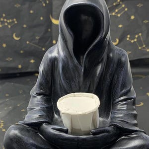 May include: A black hooded figure statue holding a white cup. The statue is in a seated position with the cup resting in its hands. The background is black with gold stars and crescent moons.