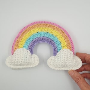 Op de afbeelding: Een gehaakte regenboog van roze, oranje, gele, groene, paarse en witte draad. De regenboog heeft twee witte gehaakte wolken aan de uiteinden.