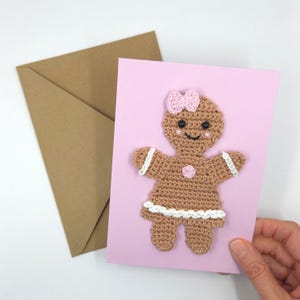 May include: A handmade greeting card featuring a crocheted gingerbread person with a pink bow and accents. The card is on a pink background, with a brown envelope in the background. The gingerbread person has a smiling face and white trim.