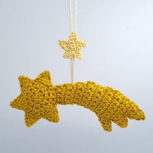 Può includere: Una stella cadente in crochet gialla con una stella gialla più piccola appesa ad essa. Le stelle sono attaccate a uno spago bianco.