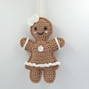 Könnte beinhalten: Gehäkelter Lebkuchenmann-Ornament mit weißer Schleife, weißer Verzierung und einem Knopf. Das braune Ornament hat ein lächelndes Gesicht mit rosigen Wangen und schwarzen Augen. Eine cremefarbene Schnur ist zum Aufhängen befestigt.