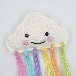 Op de afbeelding: Een witte gehaakte wolk met een glimlachend gezicht en zwarte ogen. De wolk heeft regenboogkleurige garenkwastjes die er vanaf hangen.