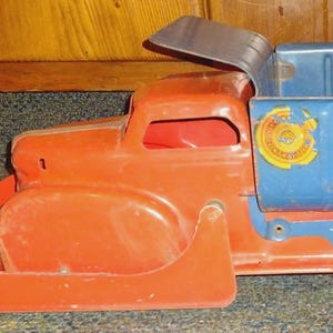 May include: A vintage metal toy dump truck featuring a bright orange cab and a blue cargo bed. The truck has a hinged blue tailgate and a circular yellow and red logo on the side of the bed. The vehicle is constructed with a sturdy frame and wheels.