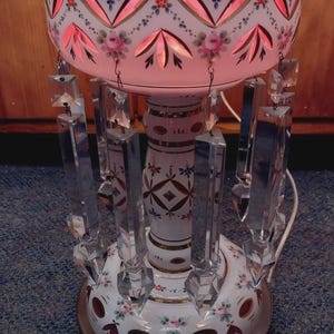 May include: A decorative table lamp with a white and pink floral design. The lamp features a bowl-shaped shade with cut-out patterns and hanging crystal prisms. The base is white with gold accents and more floral details.