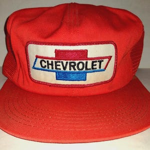May include: A red trucker hat with a white patch featuring the Chevrolet logo in blue, red, and black. The hat has a mesh back and a curved brim. The hat is a vintage style.