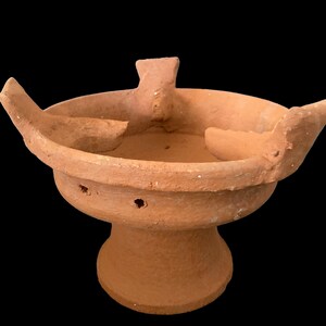 May include: A terracotta clay stove with three openings and a round base. The stove is designed for cooking over an open fire.