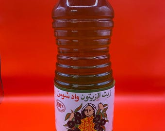May include: A clear plastic bottle of olive oil with a green cap. The label features an illustration of a woman holding olives. The bottle is filled with golden-colored oil and is labeled in Arabic and French.