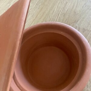 May include: A terracotta pot with a lid. The pot is round and has a wide opening. The lid is slightly ajar.