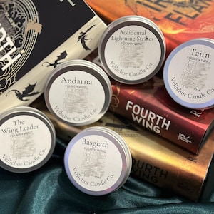 Puede incluir: Un surtido de pequeñas latas redondas de metal con etiquetas con textos como "Andarna" y "Accidental Lightning Strikes" se disponen sobre y alrededor de libros. Los libros tienen títulos como "Fourth Wing". Las latas son de Vellichor Candle Co.