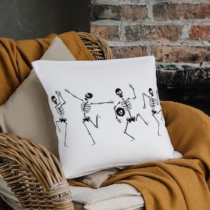Könnte beinhalten: Ein weißes Kissen mit einem schwarzen Design aus fünf tanzenden Skeletten. Das Kissen liegt auf einem Korbstuhl mit einer braunen Wolldecke.