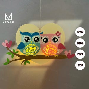 May include: Two papercraft owls with hearts and the word "Love" on their lanterns. The owls are pink and blue with black eyes and are holding lanterns with the word "Love" on them. The owls are on a branch with hearts on either side.