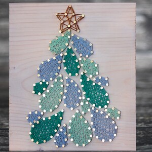 Könnte beinhalten: Ein handgefertigtes Weihnachtsbaum-String-Art-Stück auf einer Holzplatte. Der Baum ist mit blauem und grünem Faden gefertigt und mit einem goldenen Stern gekrönt. Der Baum wird mit Nägeln und Faden hergestellt und schafft eine einzigartige Weihnachtsdekoration.