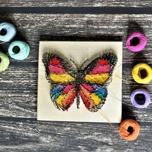 May include: A colorful string art butterfly on a wooden board. The butterfly design features a rainbow of colors, including red, yellow, and blue. Several spools of thread in various colors surround the artwork, suggesting a craft project.