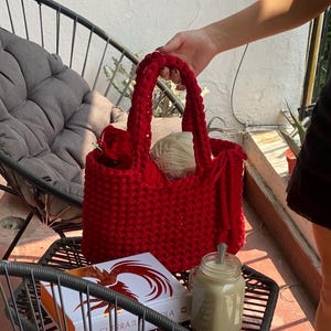 Puede incluir: Un bolso tote rojo brillante, tejido a mano, con asas gruesas, sostenido por una persona. La bolsa está llena de hilo y un ganchillo. Un libro y un tarro de café están sobre una mesa de metal negro.