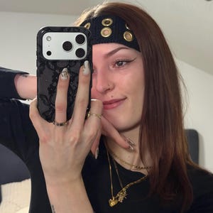 Puede incluir: Una funda de smartphone negra con un patrón de encaje texturizado, combinada con una diadema de ganchillo negra adornada con ojales metálicos de tono dorado. La funda tiene un recorte rectangular para la cámara y la diadema se lleva en la frente.