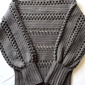 Könnte beinhalten: Ein grauer Häkelpullover mit Bootsausschnitt und langen, puffärmeligen Ärmeln. Der Pullover hat Lochmusterdetails auf Brust und Ärmeln sowie gerippte Bündchen und Saum. Das Kleidungsstück wird auf einer weißen Oberfläche präsentiert.