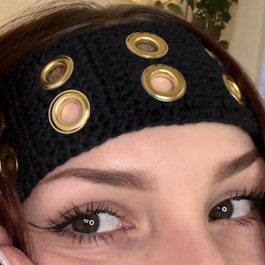 May include: Black crocheted headband adorned with multiple gold-colored eyelets. The headband is worn across the forehead. The image shows a close-up of the headband and the person's eyes.