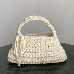 Puede incluir: Bolso de mano de ganchillo color crema con asa trenzada y base rectangular. El bolso presenta un patrón de puntadas texturizadas y un detalle de borla en un lado. El asa es arqueada y el bolso está sobre un fondo neutro.