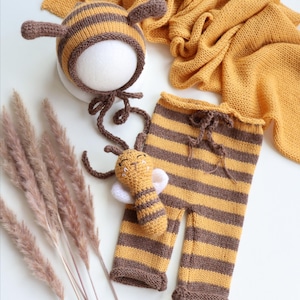 Può includere: Un set di accessori per la fotografia di neonati che comprende un cappello da ape lavorato a maglia, pantaloni e un piccolo giocattolo a forma di ape. Gli accessori sono gialli e marroni con strisce nere. Il set include anche una coperta lavorata a maglia marrone e gialla.