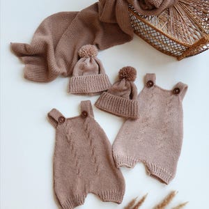 May include: A set of two knitted rompers and two matching beanies in a dusty rose color. Each romper features a cable knit pattern on the front and two brown buttons at the shoulder straps. The beanies are topped with small matching pom-poms.