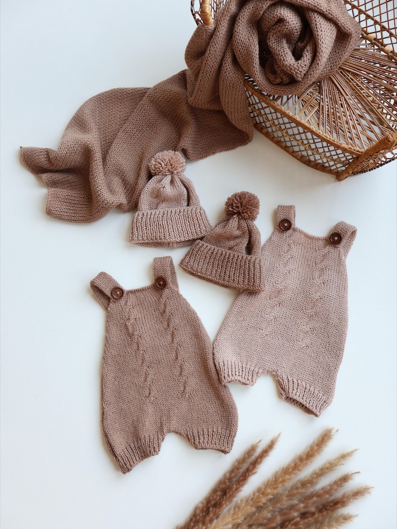 May include: A set of two knitted rompers and two matching beanies in a dusty rose color. Each romper features a cable knit pattern on the front and two brown buttons at the shoulder straps. The beanies are topped with small matching pom-poms.