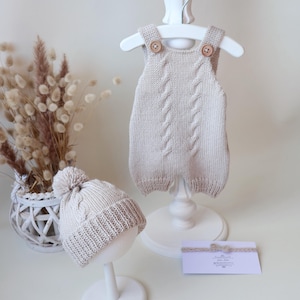 May include: A beige knitted romper with a central cable knit pattern and two wooden buttons at the shoulder straps, paired with a matching beanie featuring a pom-pom and a ribbed cuff, alongside a small rectangular card with a bow detail.