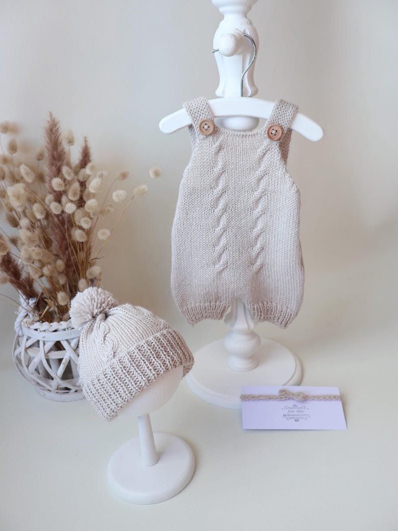 May include: A beige knitted romper with a central cable knit pattern and two wooden buttons at the shoulder straps, paired with a matching beanie featuring a pom-pom and a ribbed cuff, alongside a small rectangular card with a bow detail.