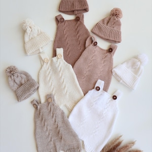 May include: A collection of knitted baby rompers and matching beanies with pom-poms. The rompers feature cable knit patterns and wooden buttons on the straps. The set includes items in solid shades of cream, white, and light brown, arranged together.