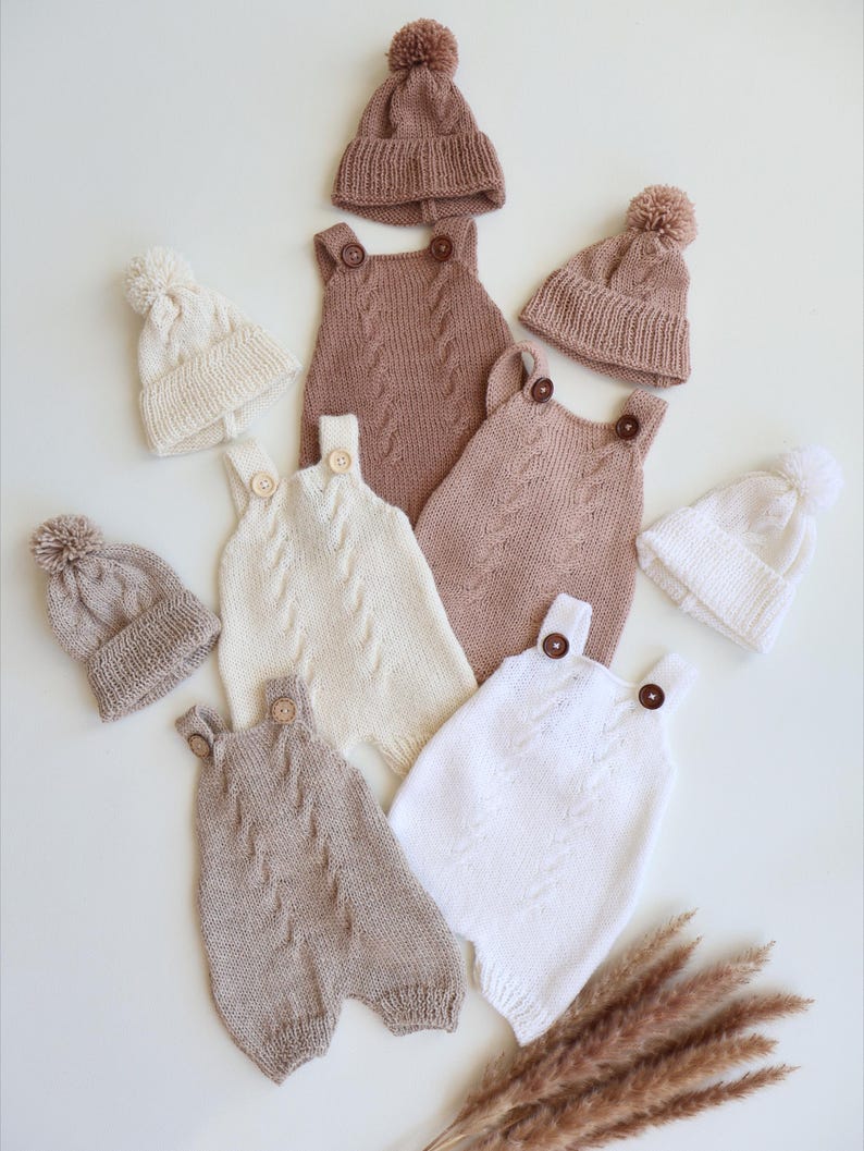 May include: A collection of knitted baby rompers and matching beanies with pom-poms. The rompers feature cable knit patterns and wooden buttons on the straps. The set includes items in solid shades of cream, white, and light brown, arranged together.