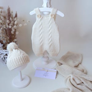 May include: A cream-colored knitted romper with two buttons at the shoulders, a matching beanie with a pom-pom, and a lightweight knit blanket. The romper features a vertical cable knit pattern down the center and ribbed leg openings.