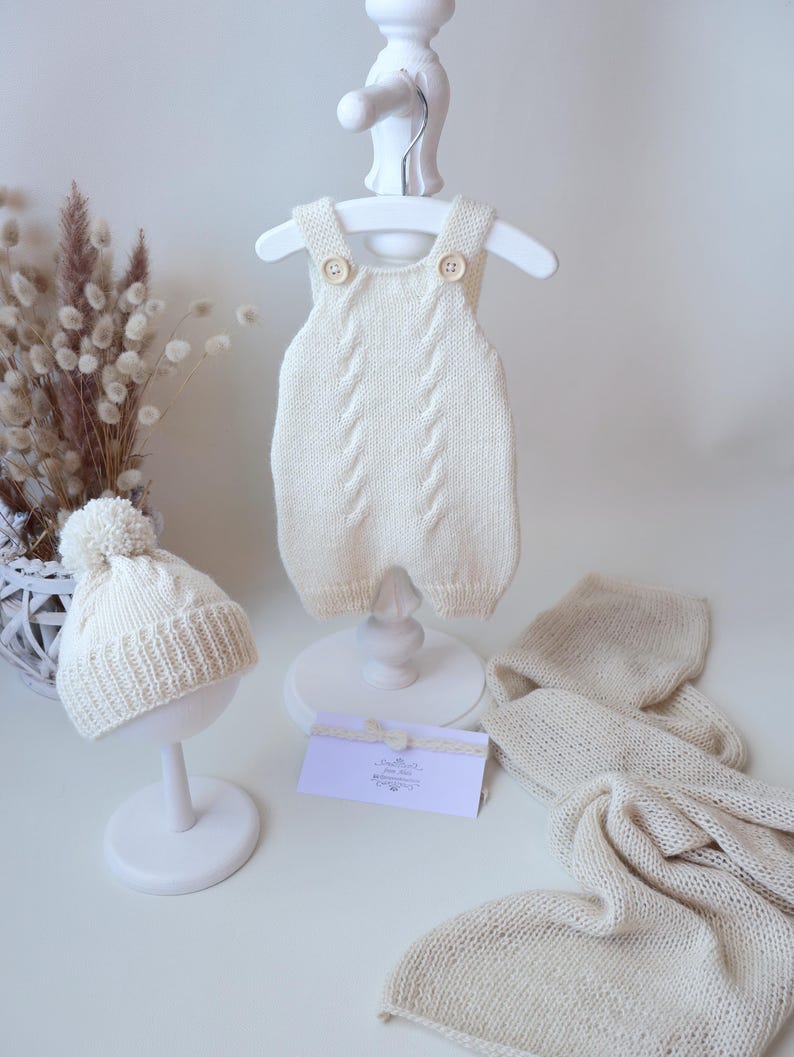 May include: A cream-colored knitted romper with two buttons at the shoulders, a matching beanie with a pom-pom, and a lightweight knit blanket. The romper features a vertical cable knit pattern down the center and ribbed leg openings.
