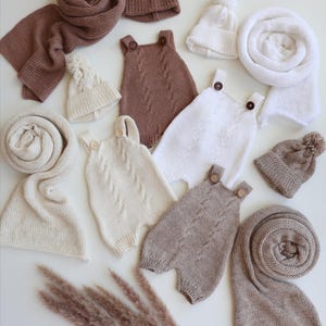 May include: A collection of knitted baby clothing including three rompers with button straps and cable knit patterns, four beanies with pom-poms, and three long scarves in shades of brown, cream, and white, arranged on a plain surface.