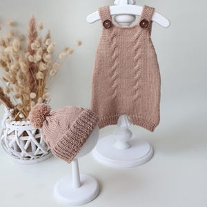 May include: A light brown knitted romper and a matching beanie with a pom-pom. The romper features a cable knit pattern on the front and two dark brown buttons on the shoulder straps. The beanie has a ribbed cuff and a cable knit design.