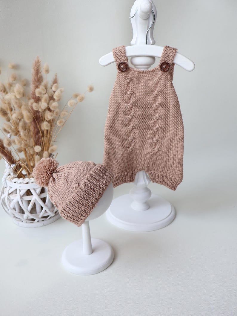 May include: A light brown knitted romper and a matching beanie with a pom-pom. The romper features a cable knit pattern on the front and two dark brown buttons on the shoulder straps. The beanie has a ribbed cuff and a cable knit design.