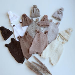 May include: A collection of knitted baby rompers and matching beanies with pom-poms in various neutral shades including white, cream, beige, tan, and dark brown. Each romper features a cable knit pattern, two shoulder straps, and wooden buttons.