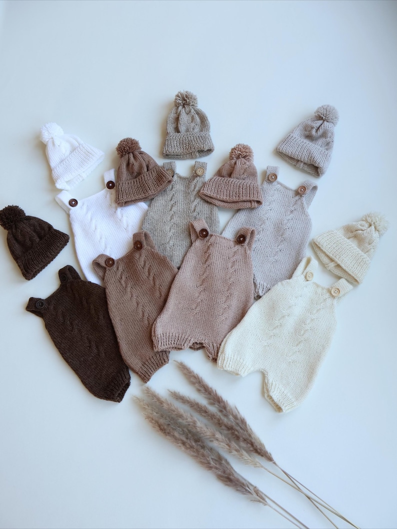 May include: A collection of knitted baby rompers and matching beanies with pom-poms in various neutral shades including white, cream, beige, tan, and dark brown. Each romper features a cable knit pattern, two shoulder straps, and wooden buttons.