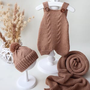 May include: A knitted set in a solid tan color consisting of a sleeveless romper with two brown buttons, a beanie with a pom-pom, and a long scarf. The romper features a vertical cable knit pattern. The text "Set #6" is visible in the bottom left.