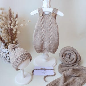 May include: A beige knitted baby romper with cable knit details and two wooden buttons, a matching pom-pom beanie, and a long knitted scarf. A small white rectangular card with text is placed in the center between the items.