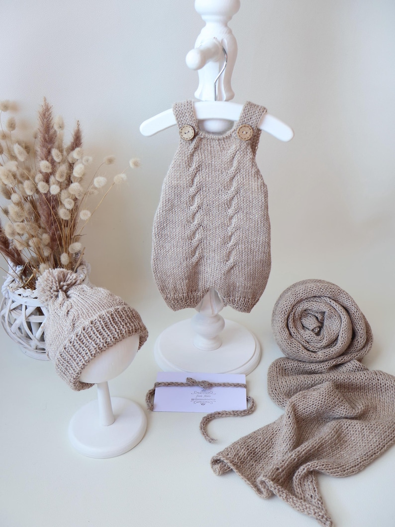 May include: A beige knitted baby romper with cable knit details and two wooden buttons, a matching pom-pom beanie, and a long knitted scarf. A small white rectangular card with text is placed in the center between the items.