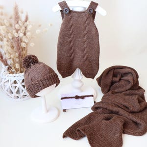May include: A brown knitted set consisting of a sleeveless romper with two buttons at the shoulders, a beanie with a pom-pom, a long scarf, and a thin headband with a small bow. Each piece features a matching cable knit pattern.
