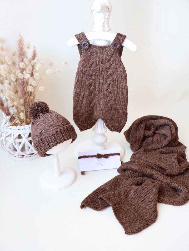 May include: A brown knitted set consisting of a sleeveless romper with two buttons at the shoulders, a beanie with a pom-pom, a long scarf, and a thin headband with a small bow. Each piece features a matching cable knit pattern.