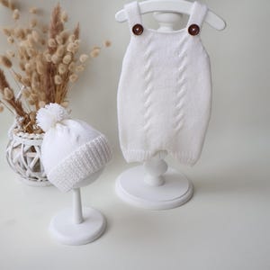 May include: A white knitted romper featuring two brown buttons on the shoulder straps and a vertical cable knit pattern, paired with a matching white knitted beanie topped with a pom-pom and finished with a ribbed cuff edge detail.