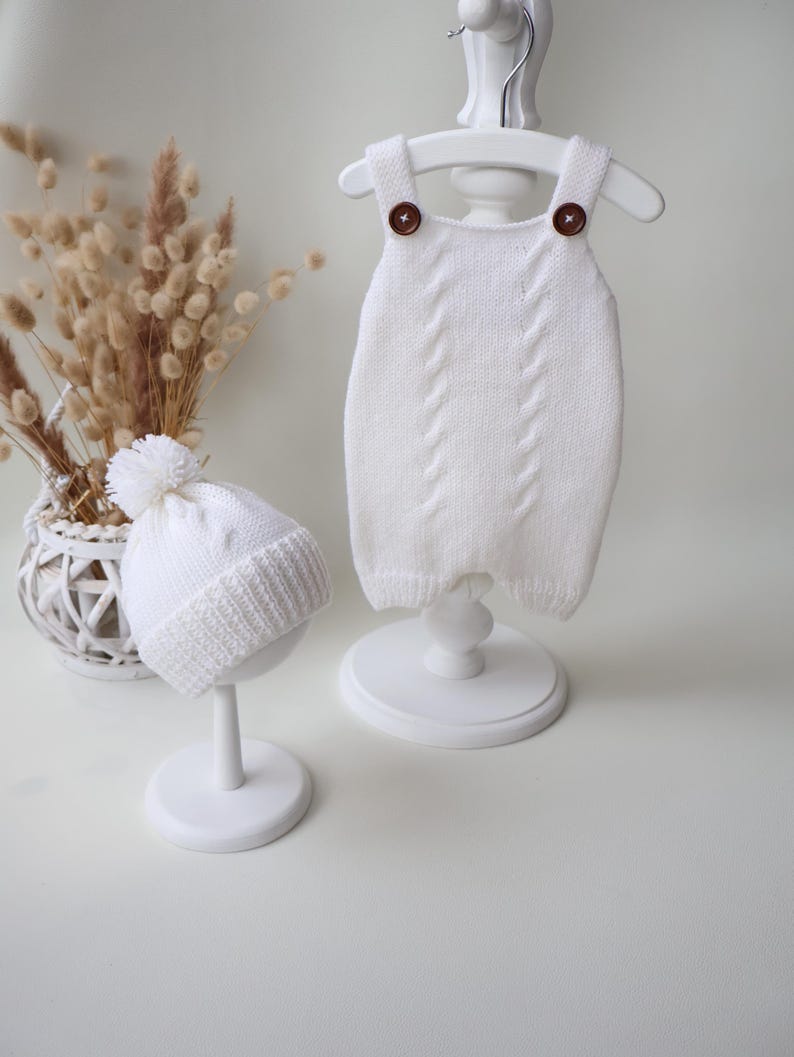 May include: A white knitted romper featuring two brown buttons on the shoulder straps and a vertical cable knit pattern, paired with a matching white knitted beanie topped with a pom-pom and finished with a ribbed cuff edge detail.