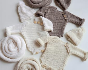 May include: An assortment of knitted baby clothing in neutral tones. The image features several cream and brown onesies with wooden buttons, matching beanies with pom-poms, and rolled blankets. The items are arranged on a white surface.