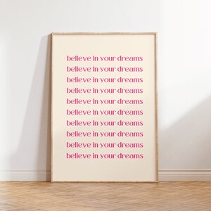 May include: A framed print with the text "believe in your dreams" repeated ten times in a pink font on a cream background.