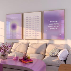 May include: Three framed motivational posters with purple and white text on a gradient background. The posters are hanging on a white wall above a beige sofa. The left poster says "Believe in your dreams". The middle poster says "I can do everything that I set my mind to".