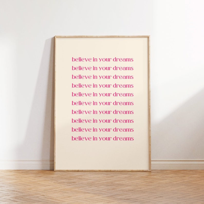 May include: A framed print with the text "believe in your dreams" repeated ten times in a pink font on a cream background.