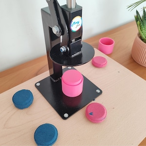 Op de afbeelding: Een zwarte knopenmaker machine met een roze knop en verschillende blauwe en roze knoppen op een houten oppervlak. Een kleine roze beker en een potplant zijn ook zichtbaar.