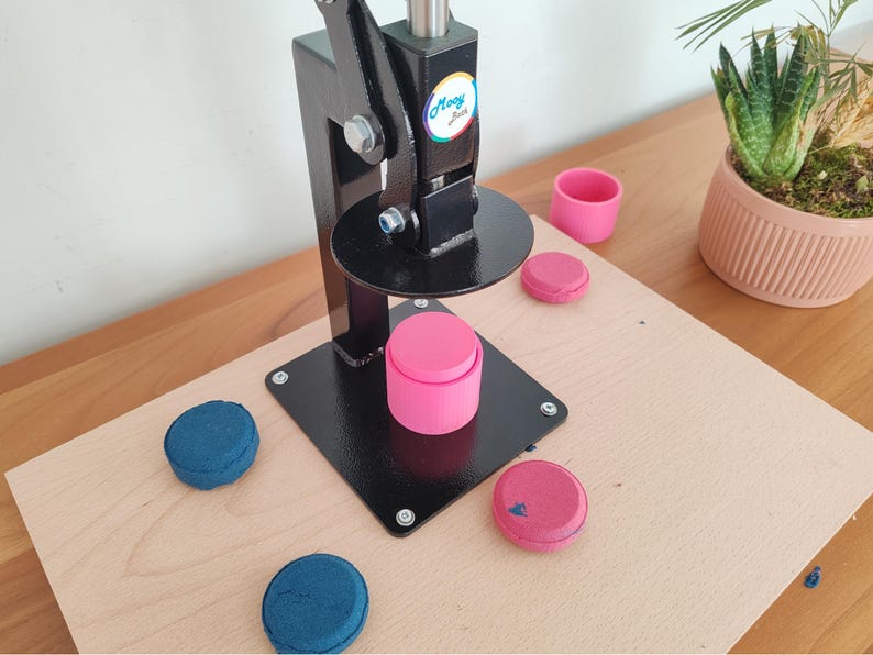 Op de afbeelding: Een zwarte knopenmaker machine met een roze knop en verschillende blauwe en roze knoppen op een houten oppervlak. Een kleine roze beker en een potplant zijn ook zichtbaar.